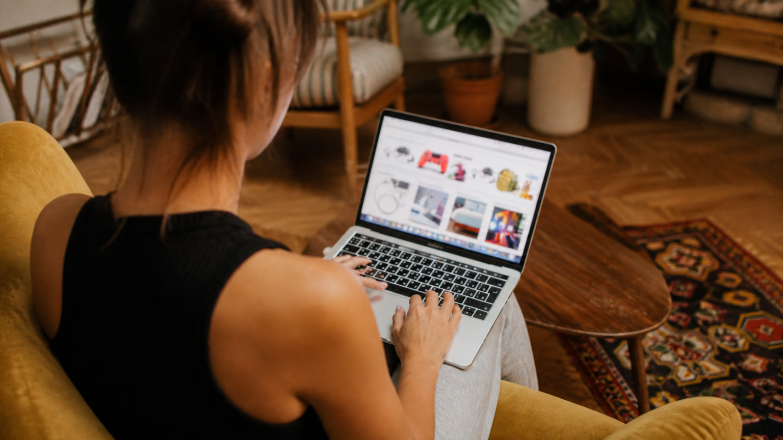 An adult seated in a living room uses a laptop displaying a blurred online shopping page.