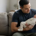 Parent holding a sleeping infant beside a closed laptop in a quiet home workspace