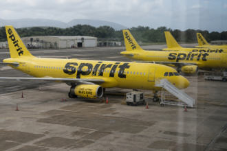 Several yellow Spirit Airlines aircraft sit on an airport apron with mobile stairs, service equipment, traffic cones, and maintenance buildings nearby.