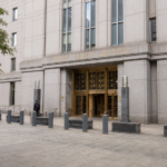 Two pedestrians walk across a tree-lined plaza outside a gray stone courthouse with brass entrance doors.