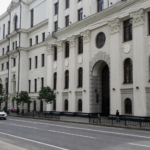 A white neoclassical government building lines a city street with passing cars and a few pedestrians.