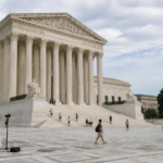 Visitors gather and walk across a broad plaza in front of a columned marble courthouse under a partly cloudy sky.