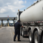 A uniformed border officer inspects a tanker truck near commercial customs lanes where other freight trucks are waiting.