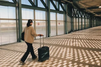 Editorial illustration of a traveler rolling a suitcase through a window-lined airport passage.
