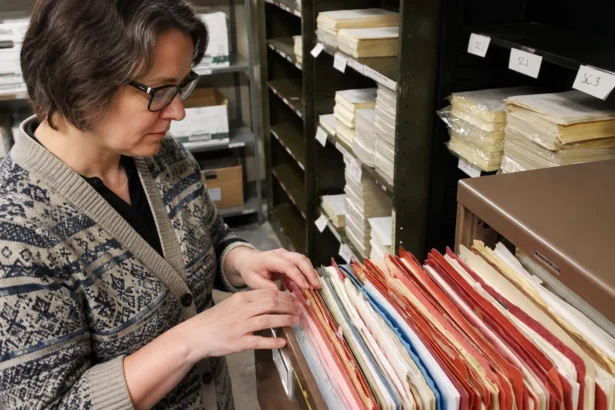 Editorial illustration of a records worker sorting folders in an open filing drawer beside archive shelves.