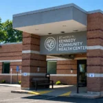 Editorial illustration of a brick community health center entrance with a canopy, bench, and accessible curb route