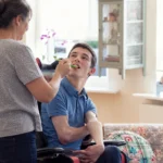 Editorial illustration viewed over a caregiver’s shoulder as she helps a younger adult in a wheelchair eat a spoonful of food at home.
